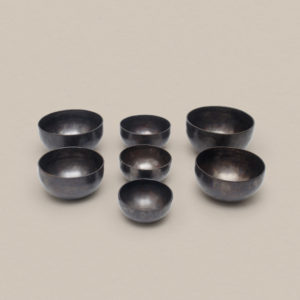 Seven hand-hammered note bowls from the Pure Bliss C4 Healing Bowl Set in a midnight black finish, arranged in varying sizes on a plain background.